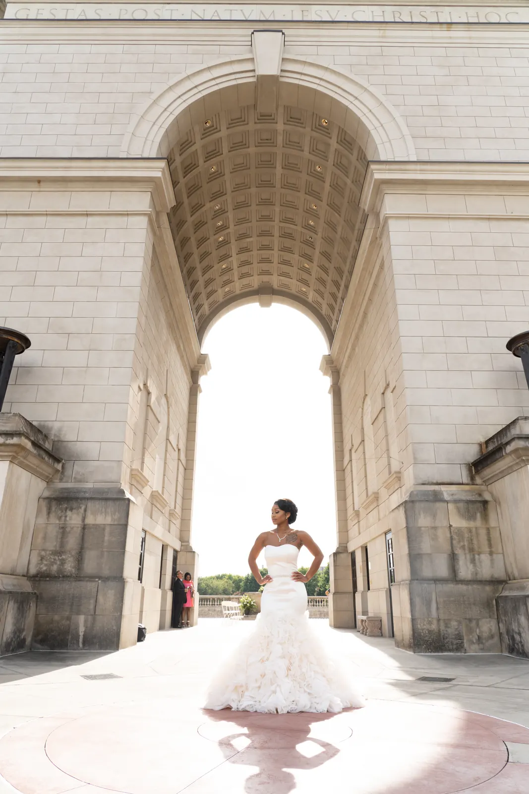 Bride in a fitted gown standing beneath a tall stone archway, photographed from behind