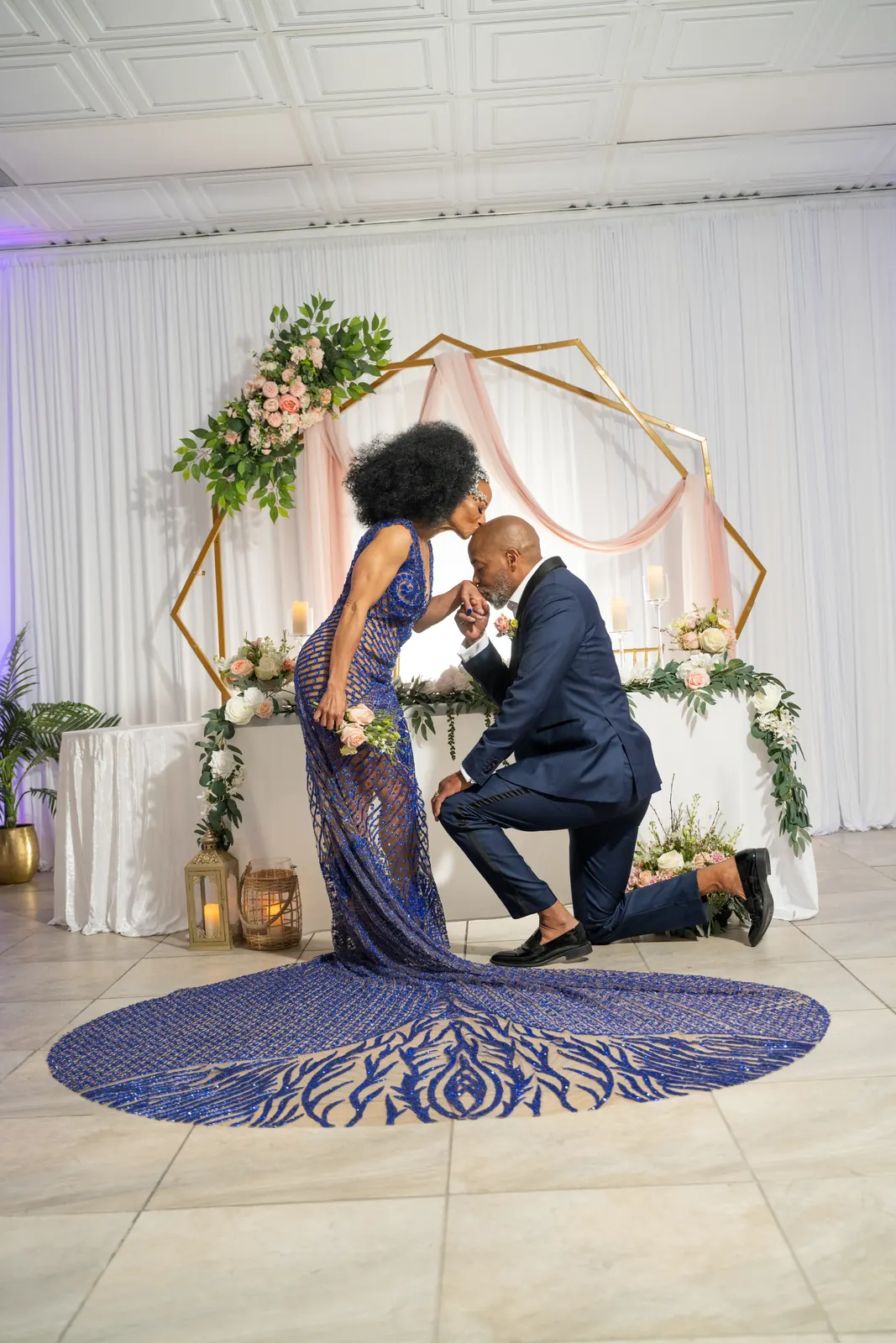 Groom kneeling before his bride in a sequined blue gown under a floral hexagon arch